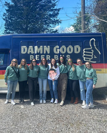 Nine friends in matching green sweatshirts smiling and posing in front of a blue-and-red beer tour bus with a large thumbs-up logo, holding an oversized face cutout on a sunny day in a gravel lot.