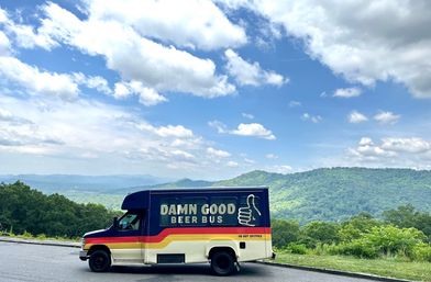 Retro-style beer bus with a thumbs-up logo parked at a scenic mountain overlook, overlooking green forested hills under a bright blue sky dotted with fluffy clouds.