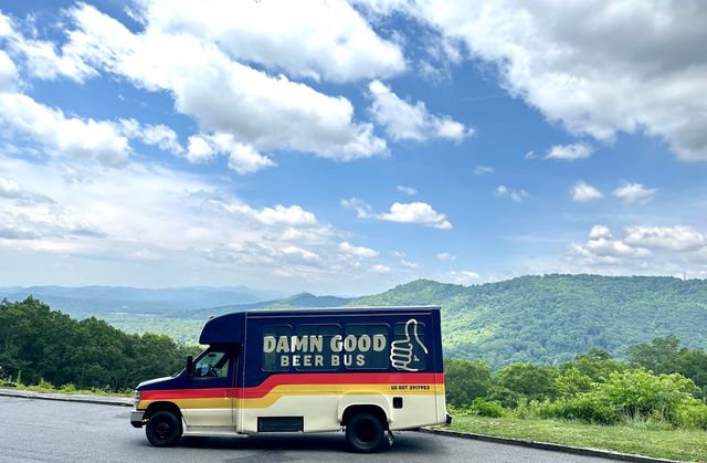 Retro-style beer bus with a thumbs-up logo parked at a scenic mountain overlook, overlooking green forested hills under a bright blue sky dotted with fluffy clouds.