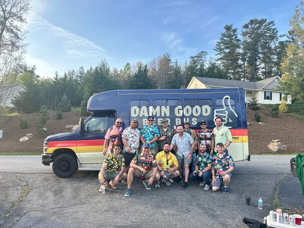 Cheerful group of friends in colorful Hawaiian shirts posing in front of a beer-themed party bus on a sunny suburban street with houses, trees, and drinks on the pavement.
