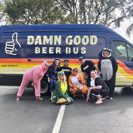 Eight adults in playful animal onesies and security shirts posing and smiling in front of a colorful beer-tour van in a parking lot.
