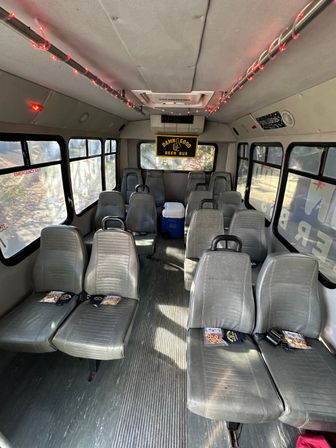 Interior of a converted shuttle/party bus with gray vinyl seats in rows, red LED ceiling lights, a yellow beer banner at the rear and a cooler in the aisle.