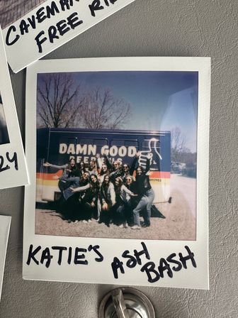 Polaroid-style group photo of a lively crew posing in front of a colorful shuttle bus with a bold slogan, bare trees and blue sky behind, handwritten party note on the white frame