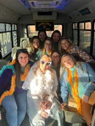 Cheerful bachelorette party inside a decorated party bus, bride-to-be in veil and heart-shaped sunglasses surrounded by smiling friends wearing orange and teal feather boas, sunlight streaming through windows.