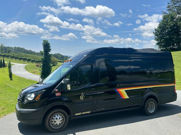 Black passenger van with colorful side stripe and thumbs-up decal parked on a winding country road beside a vineyard and rolling green hills under a bright blue sky with puffy clouds.