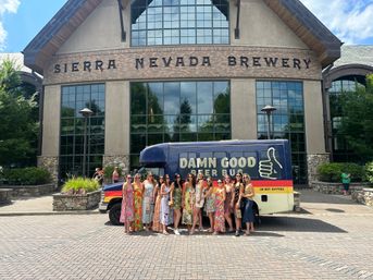 Group of women in colorful summer dresses posing by a navy beer tour bus with thumbs-up graphic outside a large craft brewery visitor center with stone and glass façade.