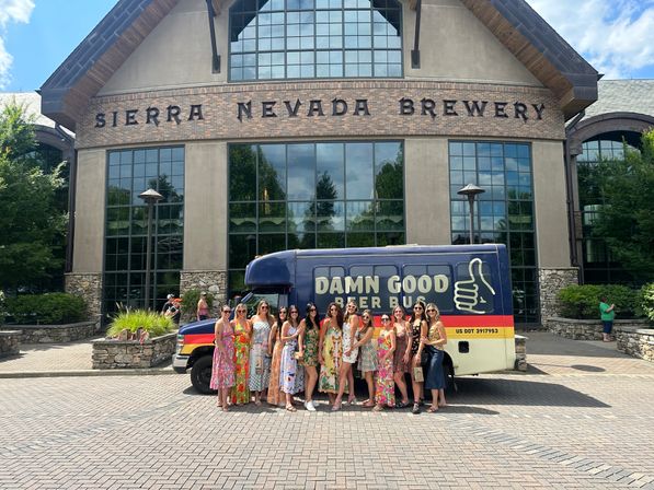 Group of women in colorful summer dresses posing by a retro beer bus parked outside a large stone-and-brick brewery entrance with tall windows and trees on a sunny day.