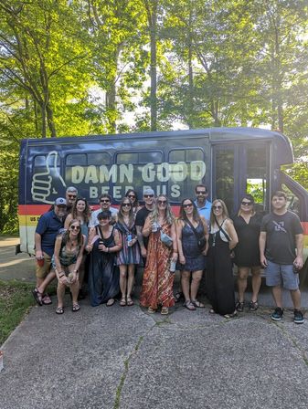 About 16 adults smiling and holding drinks, posing in front of a colorful beer-tour shuttle with a thumbs-up logo, parked on a sunlit, tree-lined driveway.