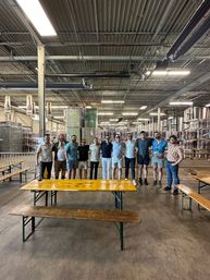 Cheerful group of people posing in a spacious brewery warehouse with towering stacks of canned beer and stainless steel tanks, yellow picnic tables in the foreground and exposed ceiling beams with bright industrial lighting.