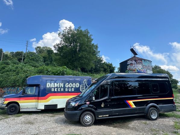 Two vans—a colorful beer-themed bus with retro stripes and a black party van—parked on a gravel lot beside a graffiti-covered silo and leafy hillside under a bright blue sky with puffy clouds.