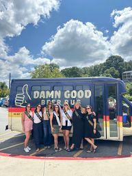 Seven friends wearing celebratory sashes pose in front of a navy beer tour party bus in a sunny parking lot beneath a bright blue sky with puffy clouds