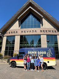 Group of friends posing by a colorful beer-tour van in front of a large alpine-style craft brewery with arched glass facade under a clear blue sky