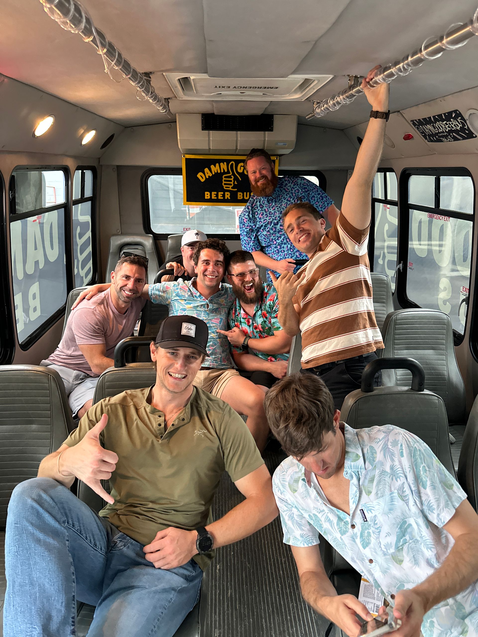 Group of eight friends laughing and posing inside a party beer bus in casual summer shirts, one making a shaka hand sign while another checks a phone