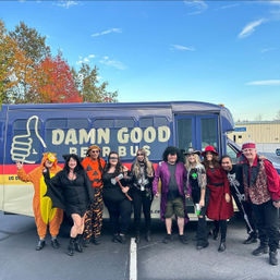 Group of adults in playful Halloween costumes posing in front of a blue party bus parked in a lot, colorful autumn trees and clear sky in the background.