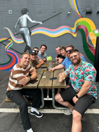 Eight friends seated at a wooden picnic table on an outdoor patio, enjoying beer flights beneath a colorful ribbon mural and painted tightrope walker