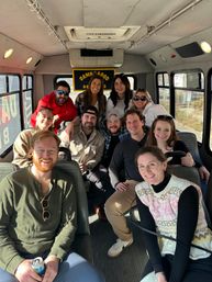 Smiling group of friends inside a sunlit party minibus on a daytime road trip, casually dressed adults seated and standing, posing for a cheerful group photo.