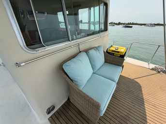 Wicker loveseat with light blue cushions on a boat deck overlooking calm marina waters and anchored boats, with teak-style decking and stainless railing visible.
