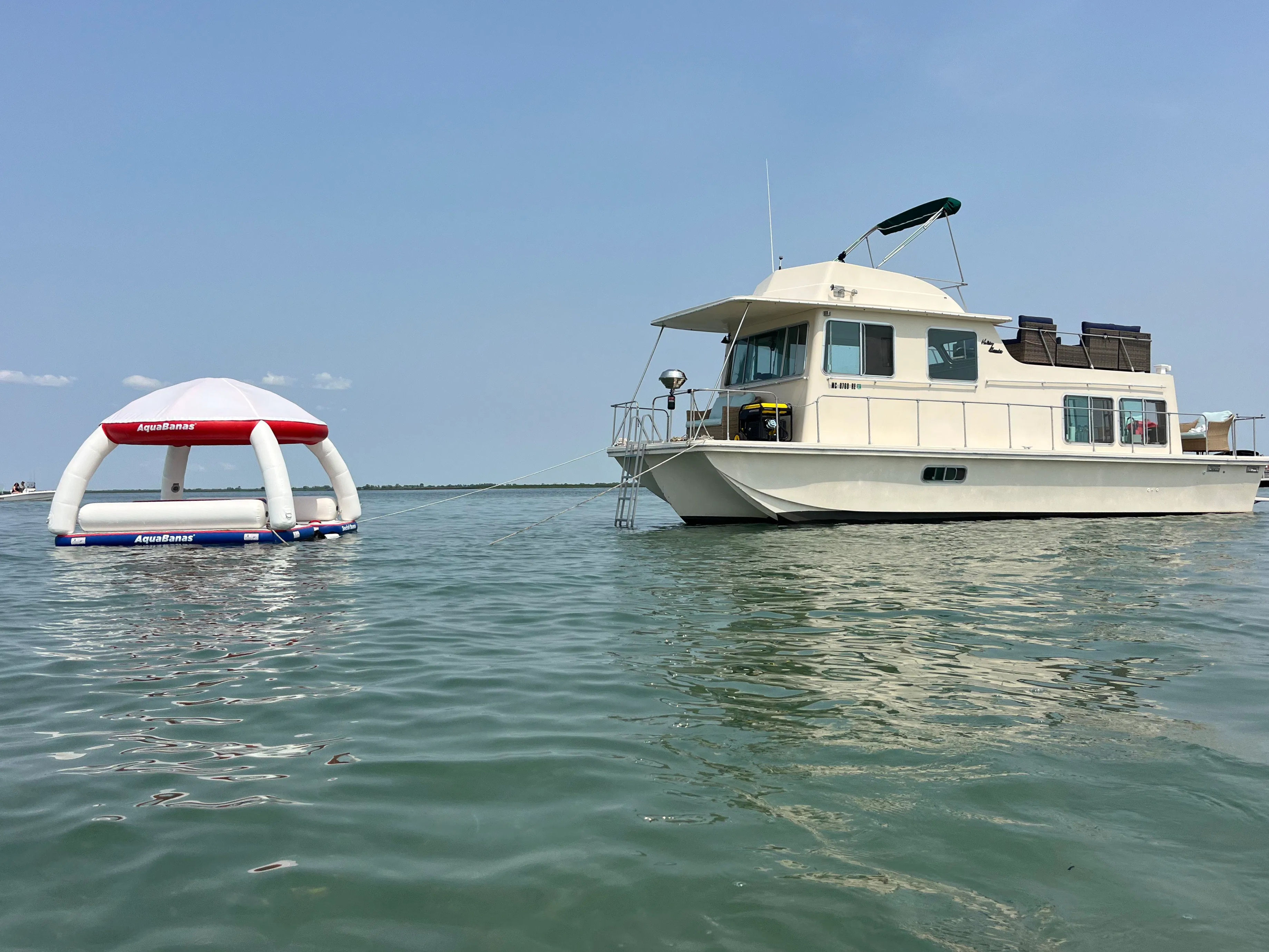 White houseboat anchored in calm coastal waters on a sunny day, with a tethered inflatable floating canopy bobbing beside it.