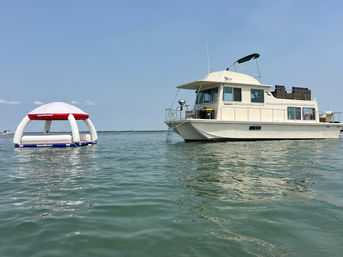 White houseboat anchored in calm coastal waters on a sunny day, with a tethered inflatable floating canopy bobbing beside it.