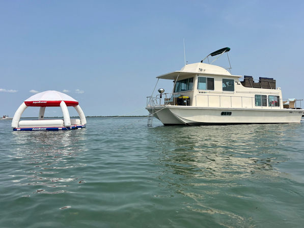 White houseboat anchored in calm coastal waters on a sunny day, with a tethered inflatable floating canopy bobbing beside it.