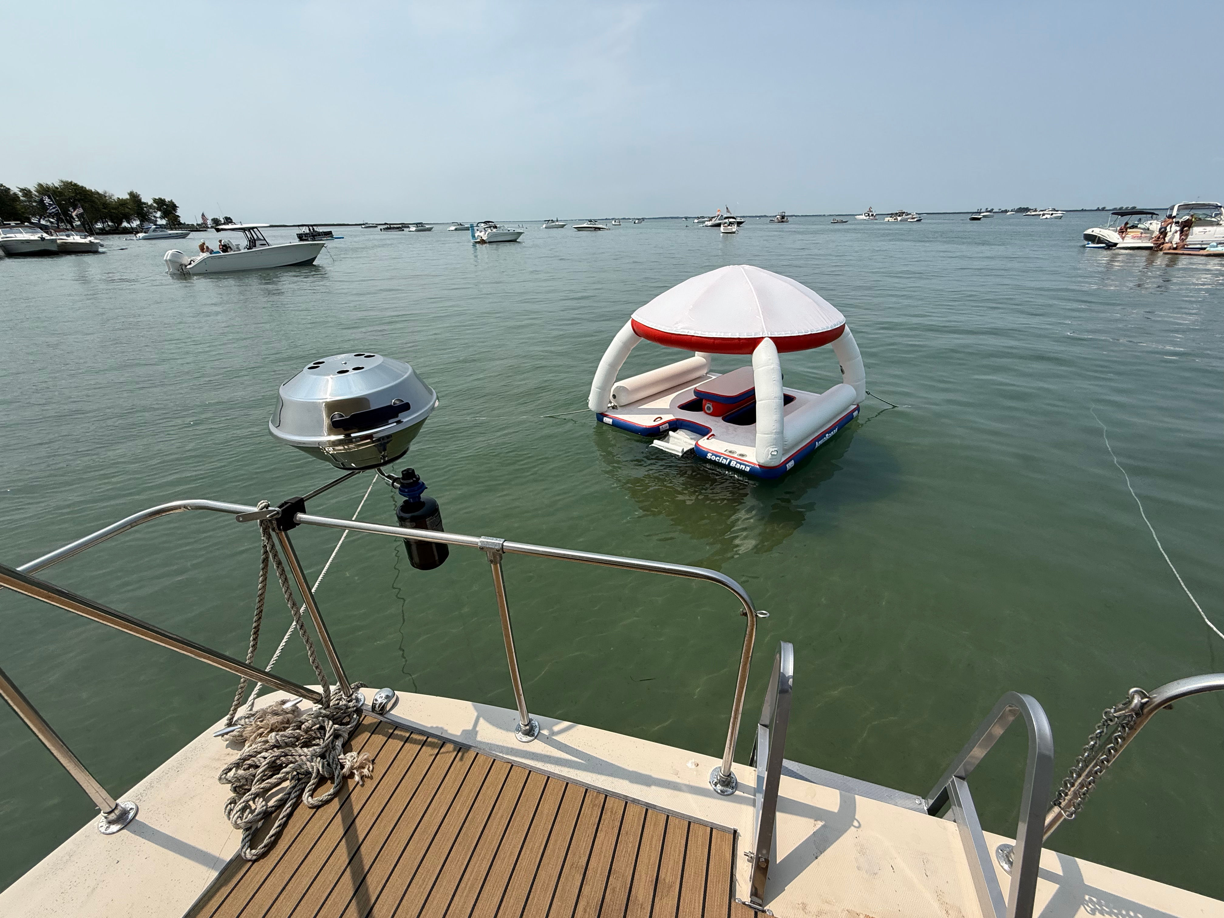 White-and-red inflatable floating lounge with canopy anchored near a boat swim platform in a busy summer bay, surrounded by multiple boats on calm green water.