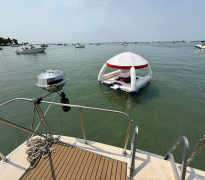 White-and-red inflatable floating lounge with canopy anchored near a boat swim platform in a busy summer bay, surrounded by multiple boats on calm green water.