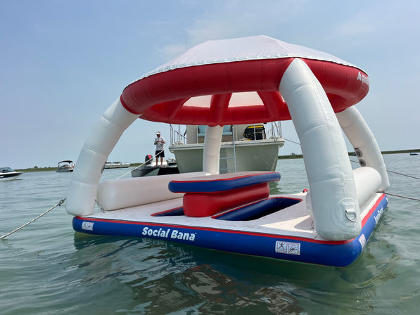 Red, white and blue inflatable floating island with shaded canopy anchored near boats in calm coastal waters on a sunny day