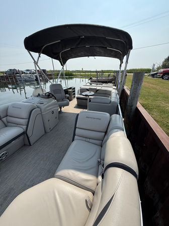 Pontoon boat with beige leather seating and black bimini canopy moored at a calm marina dock on a sunny day, ready for cruising.