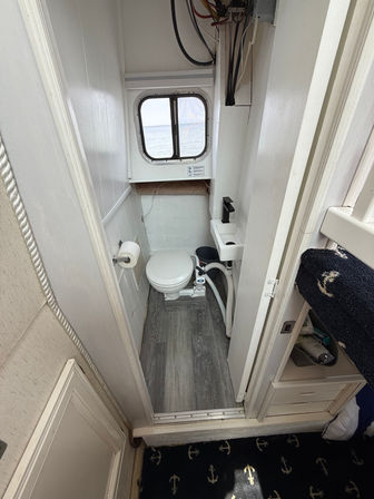 Compact boat head with white marine toilet and small sink with matte black faucet, porthole window overlooking calm water, gray wood-look floor, toilet paper holder and narrow white cabin walls.