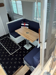 Cozy nautical dining nook inside a houseboat with navy bench cushions, striped pillow with red anchor, wooden pedestal table, and anchor-pattern carpet, windows showing turquoise water outside.