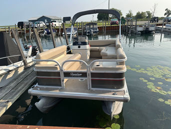 Beige-seated pontoon boat with bimini top docked at a small lake marina, calm water with lily pads and wooden piers nearby