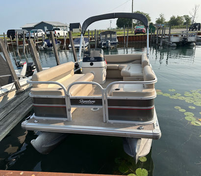 Beige-seated pontoon boat with bimini top docked at a small lake marina, calm water with lily pads and wooden piers nearby