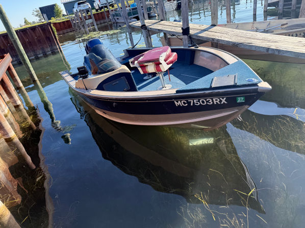 Small blue motorboat moored at a wooden dock in a sunny marina, red cushioned seat, clear shallow water with visible seagrass and dock pilings.