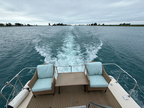 View from a boat stern with two wicker lounge chairs and a small table, turquoise wake streaming across a calm lake channel toward a distant tree-lined shore under a cloudy sky.