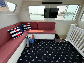 Cozy boat cabin lounge with red cushioned L-shaped seating, navy anchor-patterned carpet and striped anchor throw pillows, wall-mounted TV and large windows with marina views.