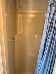 Compact beige fiberglass shower stall in a bathroom with built-in seat, chrome faucet knobs and small showerhead, partially covered by a light blue patterned shower curtain.