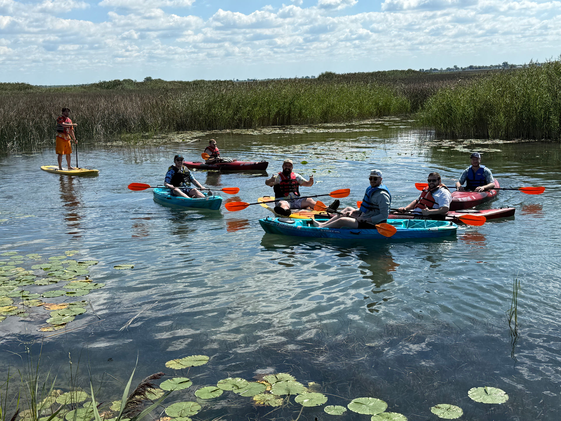 Group of paddlers in colorful kayaks and one on a paddleboard navigating a sunny freshwater marsh dotted with lily pads, tall reeds, and a blue sky with scattered clouds.