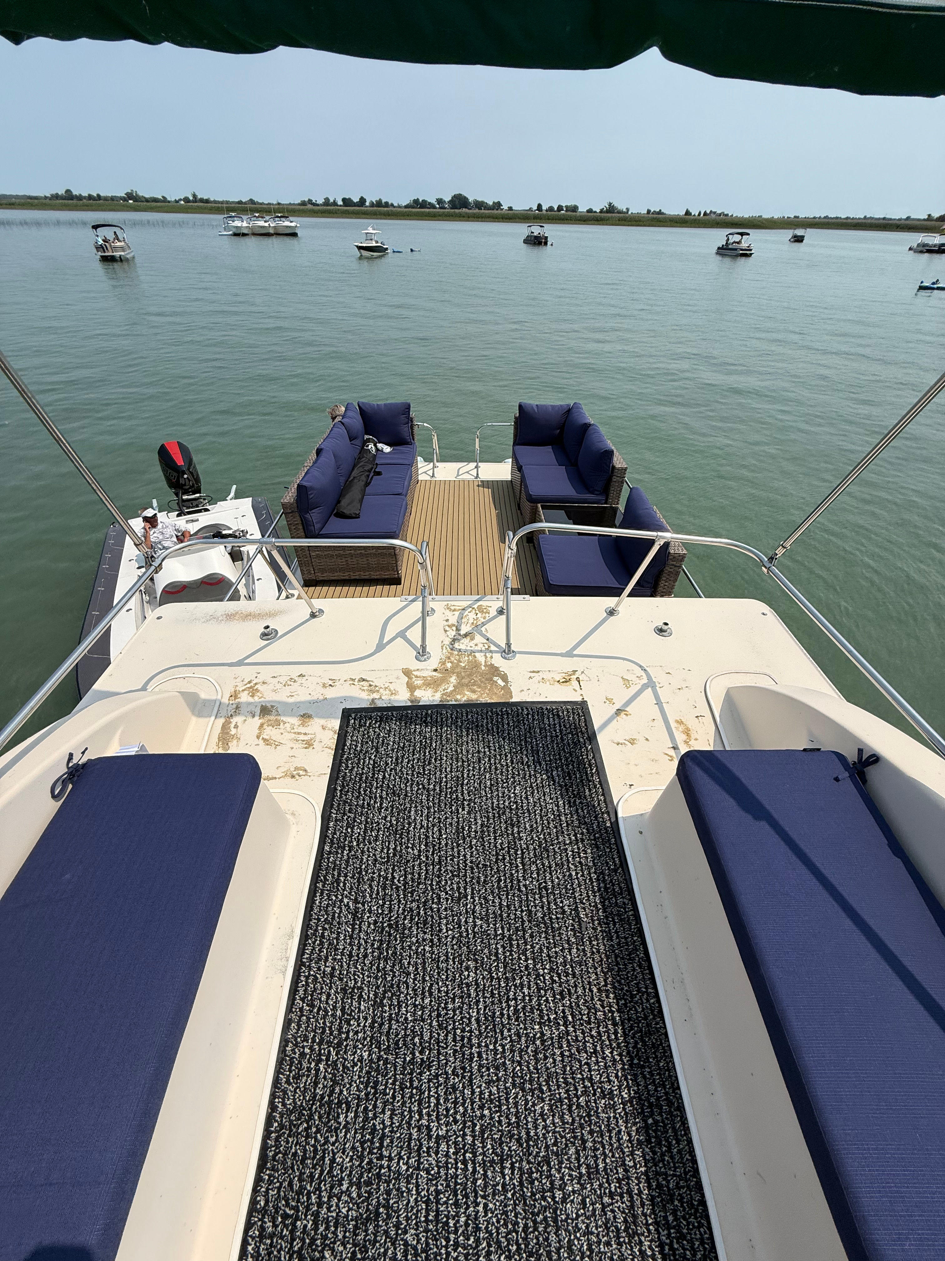 Pontoon boat stern deck with navy-cushioned seats and wicker sofas, looking out over a calm lake dotted with anchored boats and a distant shoreline on a clear summer day.