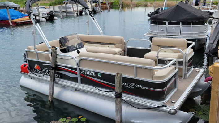 Beige-and-gray pontoon boat with open deck seating moored at a wooden dock in a calm marina, surrounded by neighboring boats and lily pads on the water.