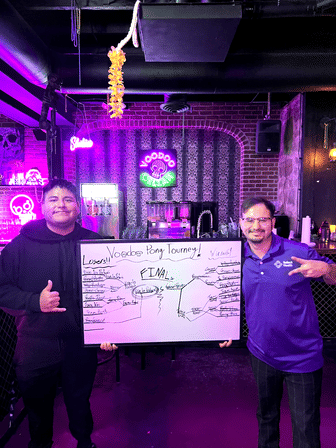 Two smiling people in a purple neon-lit bar hold a whiteboard with a beer pong tournament bracket, posing in front of a brick wall with neon skull artwork and a drink station.