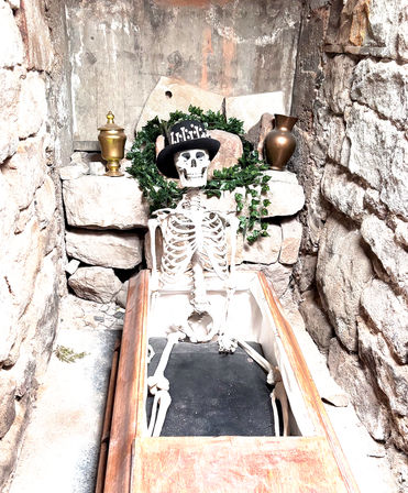 Playful skeleton sitting in an open wooden coffin wearing a black skull-adorned top hat, framed by a green wreath and brass urns against rough stone walls in a tomb-like alcove.