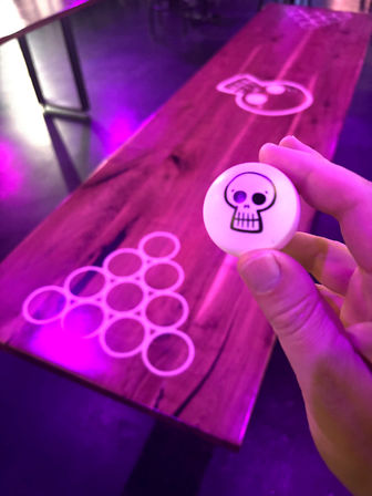 Hand holding a white ping-pong ball with a black skull logo above a wooden beer-pong table and plastic cup rack, lit by purple neon in an indoor bar/party venue.
