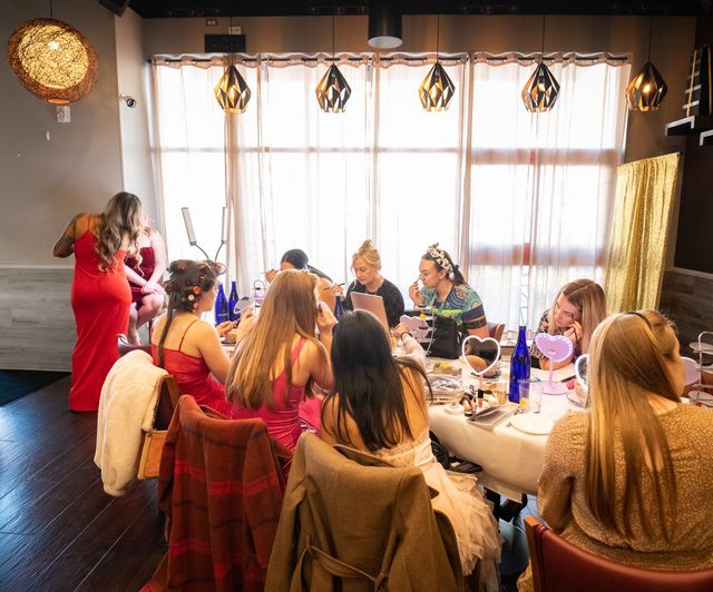Bridal-party getting-ready scene in a sunlit studio: a group of women and bridesmaids around a table applying makeup with tabletop mirrors, beauty products, rollers and hanging pendant lights