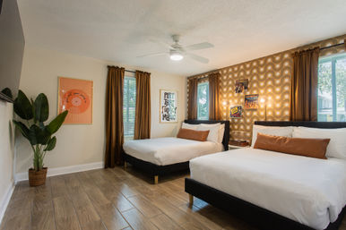 Bright retro-inspired guest bedroom with two queen beds, white bedding and tan bolster pillows, gold-brown geometric accent wall, wood-look tile floor, ceiling fan, tall windows with brown curtains and a potted plant