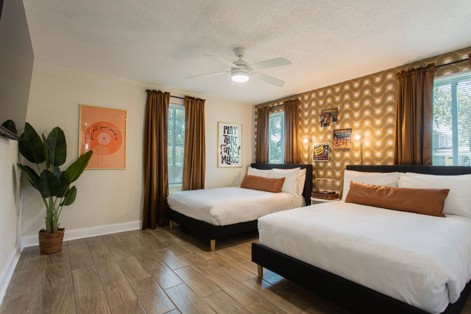 Bright retro-inspired guest bedroom with two queen beds, white bedding and tan bolster pillows, gold-brown geometric accent wall, wood-look tile floor, ceiling fan, tall windows with brown curtains and a potted plant