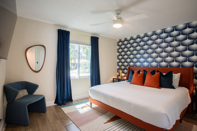 Bright modern bedroom with king-size orange-upholstered bed, navy and orange pillows, bold blue geometric accent wall, navy curtains framing a sunny window, ceiling fan, curved blue accent chair and wood-look floor.