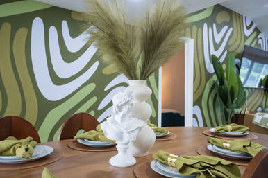 Modern dining room with wooden table set for six, green abstract mural wall, white ceramic bust and sculptural vase holding pampas grass as centerpiece, olive-green napkins and large indoor plant.