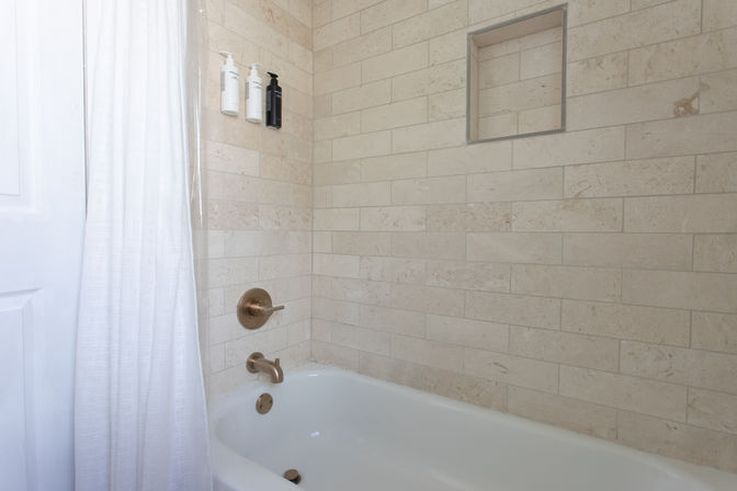 Modern bathroom bathtub with white shower curtain, beige subway-style stone tile surround, built-in niche, brushed gold faucet and wall-mounted soap dispensers