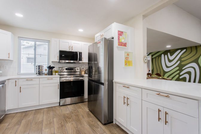 Bright modern open-concept kitchen with white shaker cabinets, brass hardware, stainless steel fridge and stove, built-in microwave, quartz counters, wood-look tile floor and a pass-through to a dining area with a green abstract accent wall.