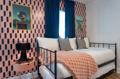 Retro-chic bedroom with black metal daybed, striped pillows, peach textured throw, pink geometric wallpaper, navy curtains and a fringe table lamp.